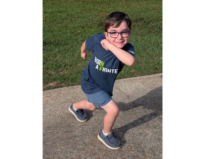 A young boy wearing glasses and a "Born A Fighter" shirt poses energetically on a sidewalk with grass in the background.