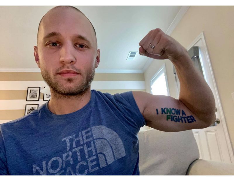 A man in a blue North Face shirt flexes his arm, displaying a tattoo or writing on his bicep that says "I KNOW A FIGHTER.