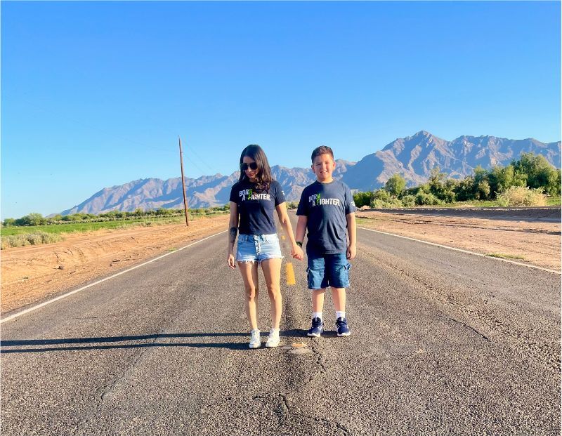 Two children wearing matching shirts stand holding hands on a rural road with mountains and clear blue sky in the background.