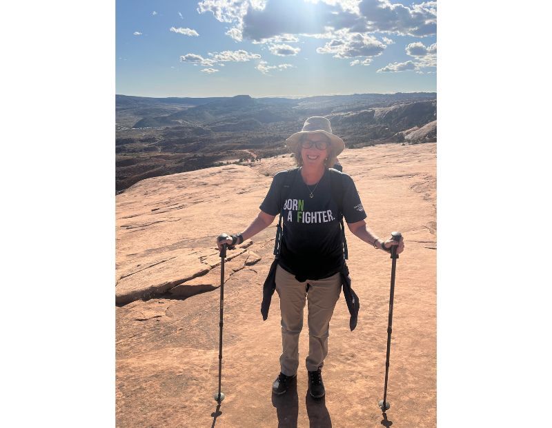 A person stands on a rocky plateau holding hiking poles, wearing sunglasses, a hat, and a shirt that reads "Born A Fighter." The sky is bright with scattered clouds.