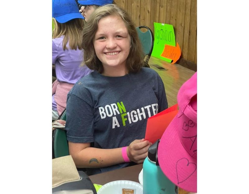 A young woman wearing a "Born a Fighter" t-shirt smiles at a table, holding a red card. Colorful hats and signs are visible in the background.