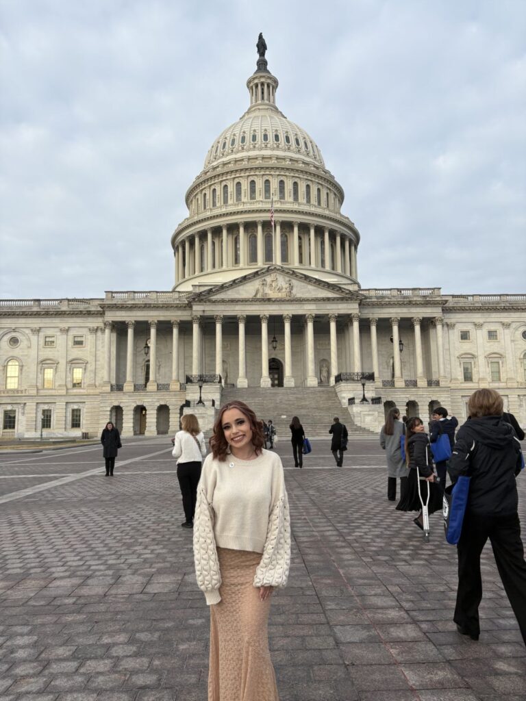 A woman stands smiling in front of the United States Capitol building with several people walking in the background on a cloudy day.