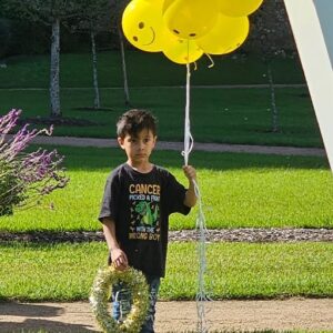 Young boy standing outdoors holding yellow balloons and a wreath, wearing a dark shirt with a cancer awareness message, with grass and trees in the background.