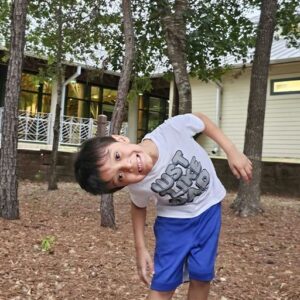 Young boy wearing a "Just Be Kind" t-shirt and blue shorts leans playfully to the side outdoors, with trees and a building in the background.