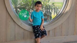 Young boy in a blue shirt and black shorts holds a green frisbee while sitting on a wooden ledge in front of a large round window with plants outside.