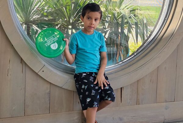 Young boy in a blue shirt and black shorts holds a green frisbee while sitting on a wooden ledge in front of a large round window with plants outside.