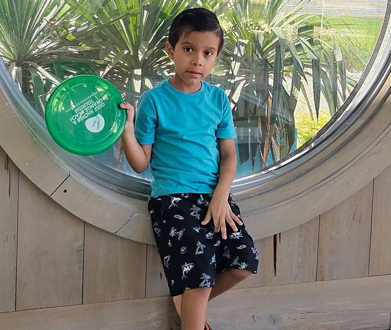 Young boy in a blue shirt and black shorts holds a green frisbee while sitting on a wooden ledge in front of a large round window with plants outside.