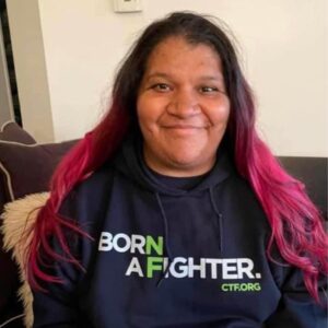 Person with long dark hair and pink ends sits on a couch, smiling, wearing a black hoodie that says "BORN A FIGHTER. CTF.ORG.