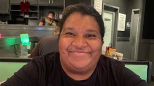 A person with dark hair and a black shirt smiles while sitting in a restaurant booth, with kitchen equipment and condiments visible in the background.