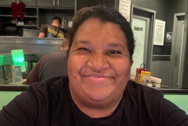 A person with dark hair and a black shirt smiles while sitting in a restaurant booth, with kitchen equipment and condiments visible in the background.