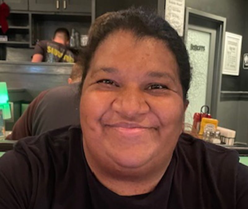 A person with dark hair and a black shirt smiles while sitting in a restaurant booth, with kitchen equipment and condiments visible in the background.