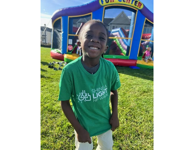 Smiling child wearing a green "Shine a Light NF Walk" T-shirt stands on grass in front of an inflatable play structure at an outdoor event.