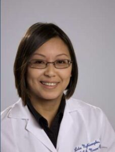 Person with short dark hair and glasses wearing a white lab coat and dark shirt, facing the camera and smiling against a plain background.