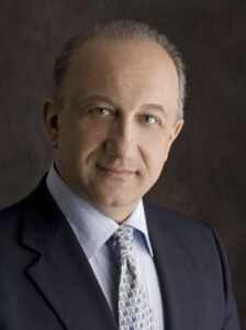 A middle-aged man in a suit and patterned tie poses for a professional portrait against a dark background.