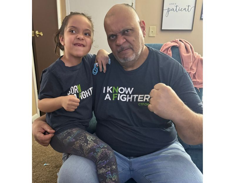 An adult and a child sit together indoors, both wearing matching shirts that say "I KNOW A FIGHTER." They are making fists and posing for the camera.