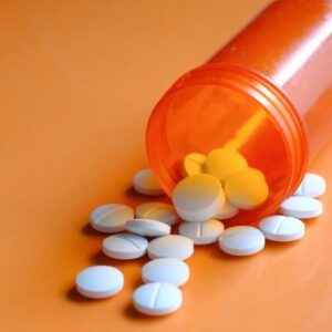 An orange prescription bottle tipped over, spilling several white round tablets onto an orange surface.