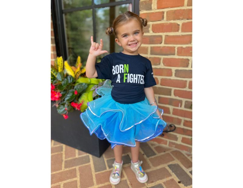 Young girl wearing a "Born a Fighter" t-shirt and blue tutu stands on a brick patio, smiling and making a hand gesture, with plants in the background.