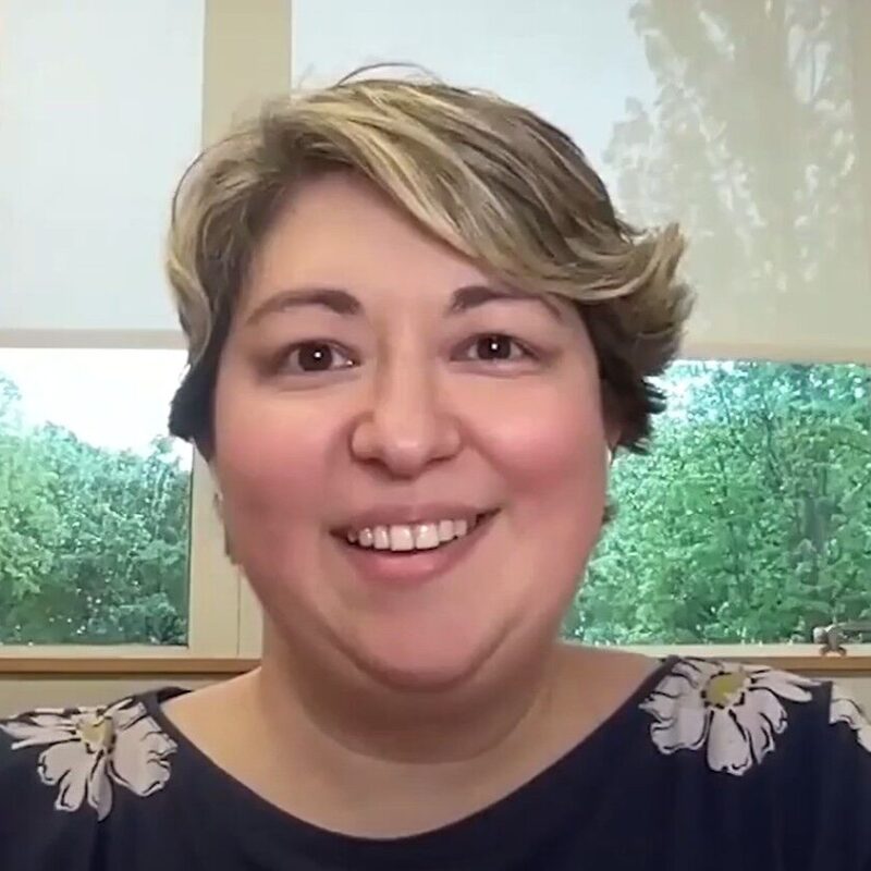 A person with short, light brown hair smiles at the camera, seated indoors in front of a window with trees visible outside.
