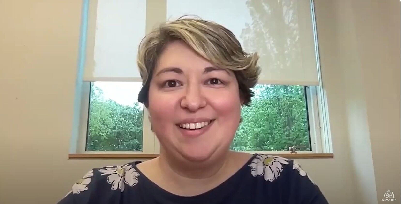 A person with short, light brown hair smiles at the camera, seated indoors in front of a window with trees visible outside.