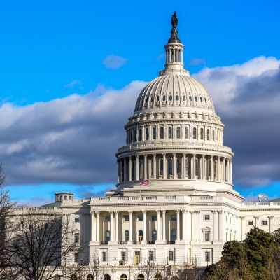 The United States Capitol building with its white dome and columns, set against a blue sky with clouds and some trees in the foreground.