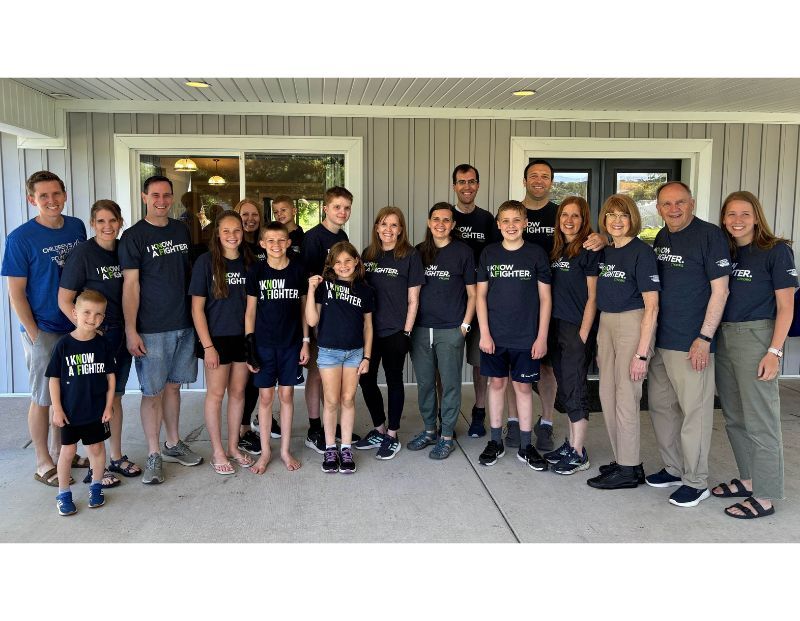 A group of people of various ages stand together outside a building, many wearing matching “I Know a Fighter” shirts and smiling at the camera.