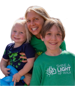 A woman poses with two smiling children, one holding a blue balloon animal. They are all wearing shirts, one with the text "SHINE A LIGHT NF WALK.
