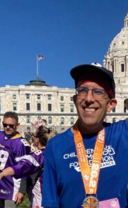 A man wearing a blue shirt and marathon medal smiles in front of a historic building, with other people and an American flag visible in the background.