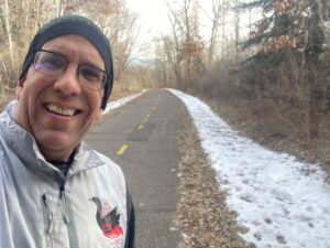 A man in a winter jacket and hat smiles for a selfie on a paved trail, with patches of snow and bare trees visible along the path.