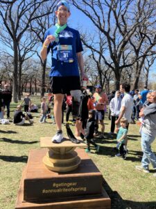 A man with a prosthetic leg stands on a pedestal holding a medal and water bottle, surrounded by people at an outdoor event. The pedestal reads "#gettingear #annualriteofspring.