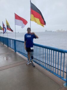 A man with a prosthetic leg stands on a riverside walkway, holding a medal. He is wearing a race bib and is surrounded by several national flags.