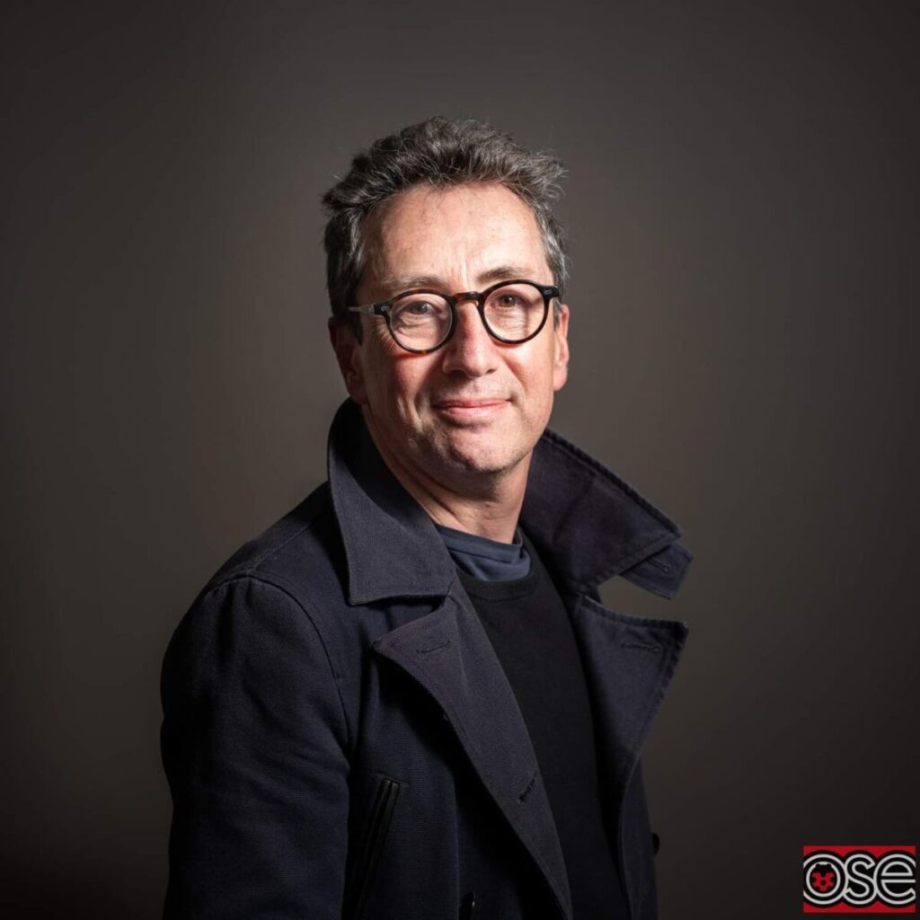 A middle-aged man with short dark hair, wearing glasses and a dark coat, stands in front of a neutral background, looking at the camera.