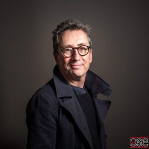 A middle-aged man with short dark hair, wearing glasses and a dark coat, stands in front of a neutral background, looking at the camera.