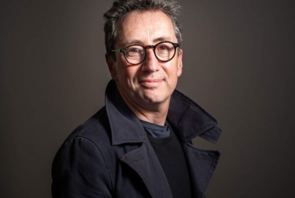 A middle-aged man with short dark hair, wearing glasses and a dark coat, stands in front of a neutral background, looking at the camera.
