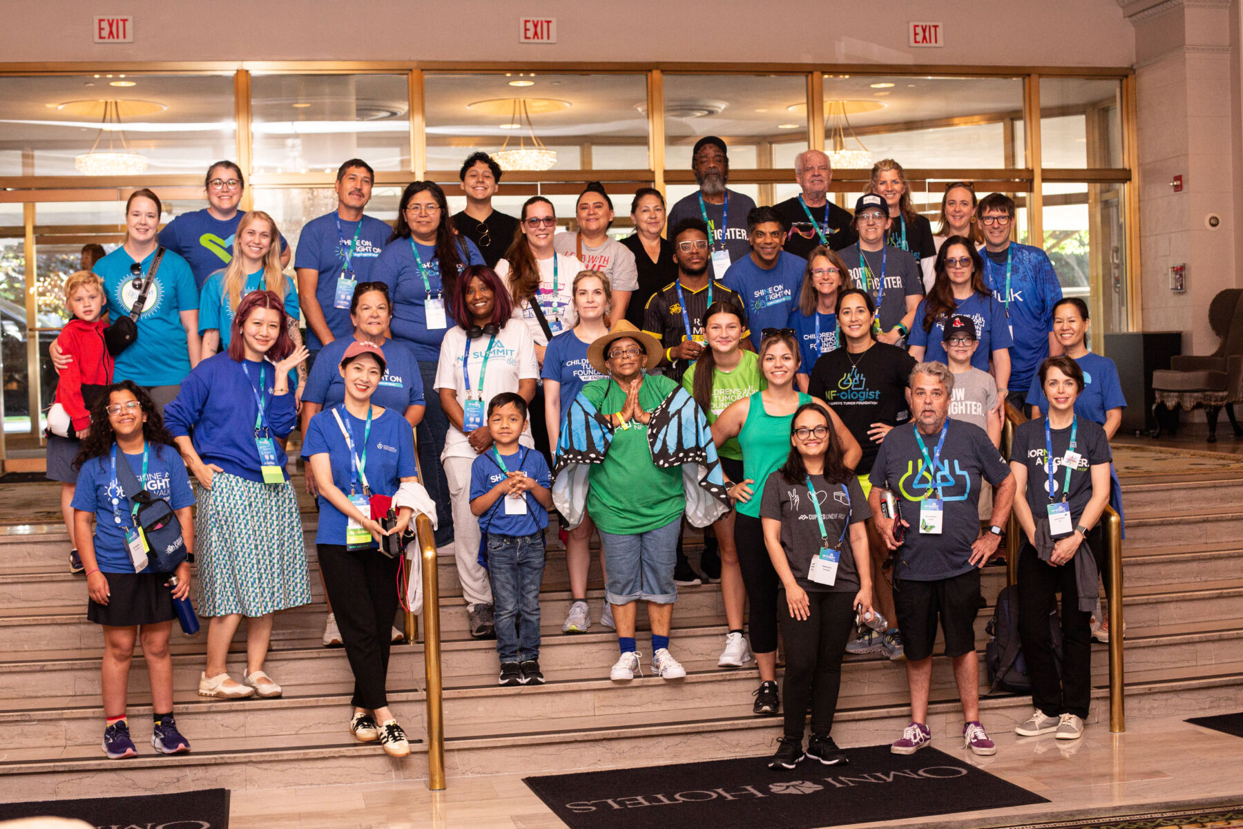 A large group of people pose together on a hotel staircase, many wearing blue shirts and name badges, for a group photo at an event or conference.