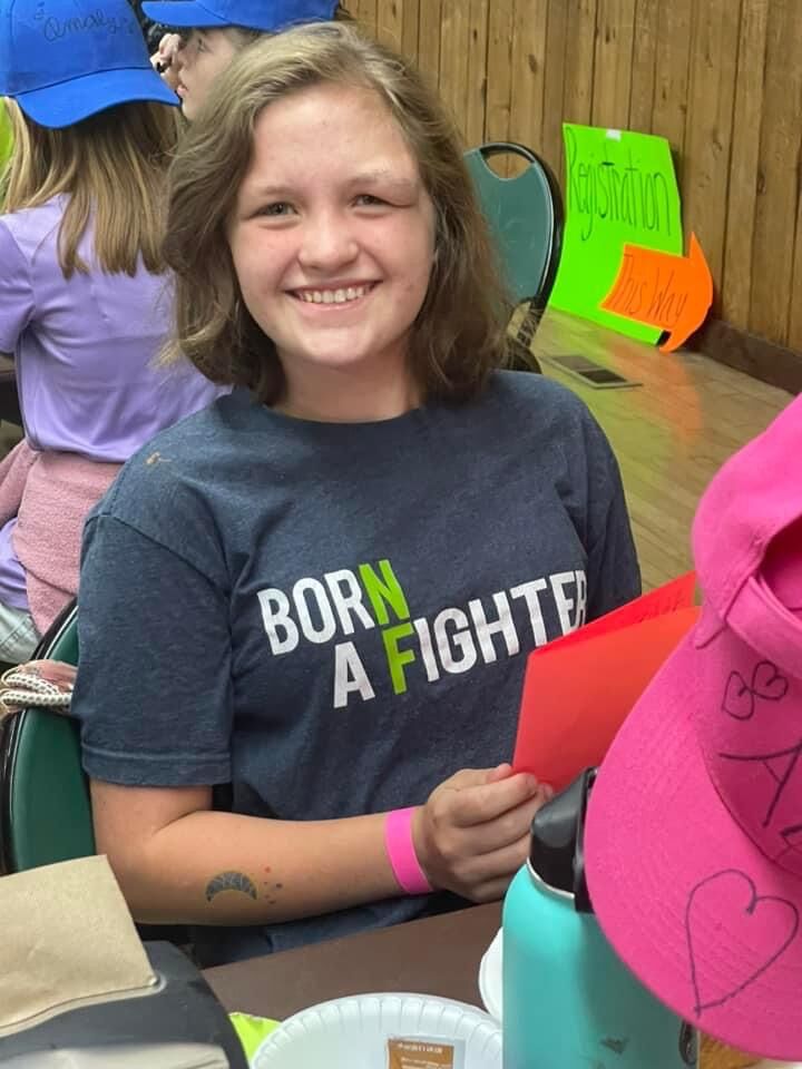 Smiling girl wearing a "Born a Fighter" shirt sits at a table holding a red paper; colorful signs and other kids are in the background.