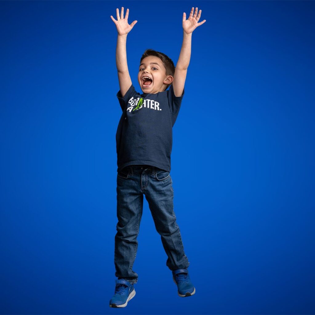 Young boy in jeans and a navy T-shirt stands against a blue background with arms raised and a joyful expression, as if jumping or celebrating.