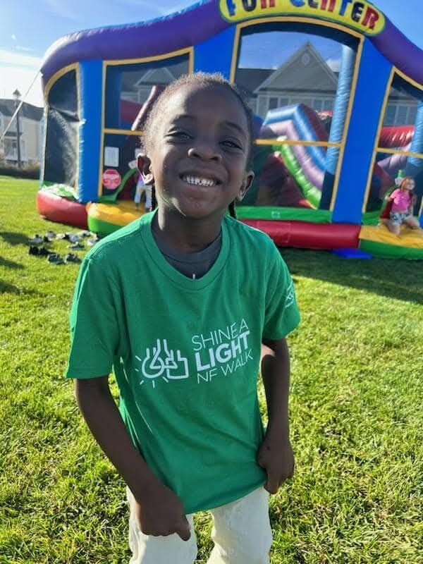 Smiling child wearing a green "Shine A Light NF Walk" T-shirt stands on grass in front of a colorful inflatable bounce house at an outdoor event.