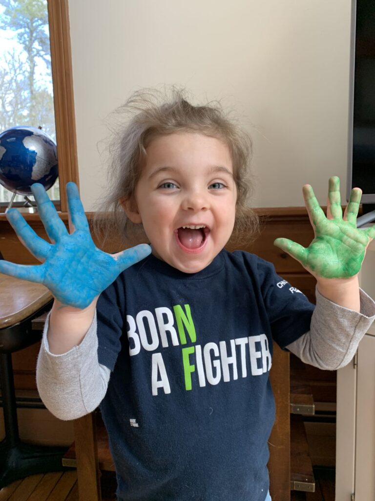 Young child smiling with both hands raised, palms covered in blue and green paint, wearing a "BORN A FIGHTER" shirt. A globe is visible in the background.