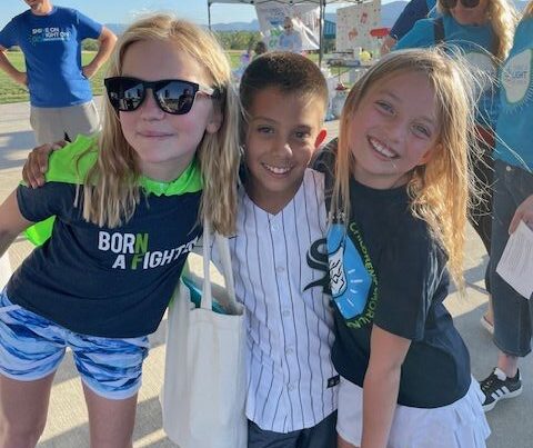 Three children stand close together smiling outdoors at an event, with adults and a tent visible in the background on a sunny day.