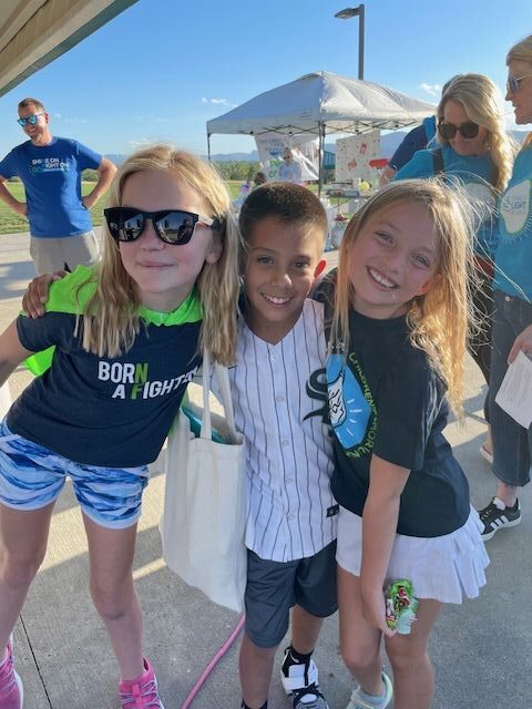Three children stand close together smiling outdoors at an event, with adults and a tent visible in the background on a sunny day.