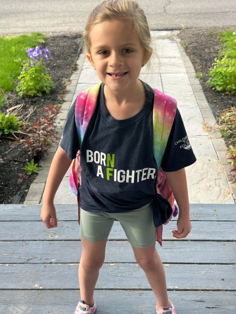 A young girl stands on a porch wearing a "Born a Fighter" t-shirt, shorts, sneakers, and a colorful backpack. She is smiling at the camera.
