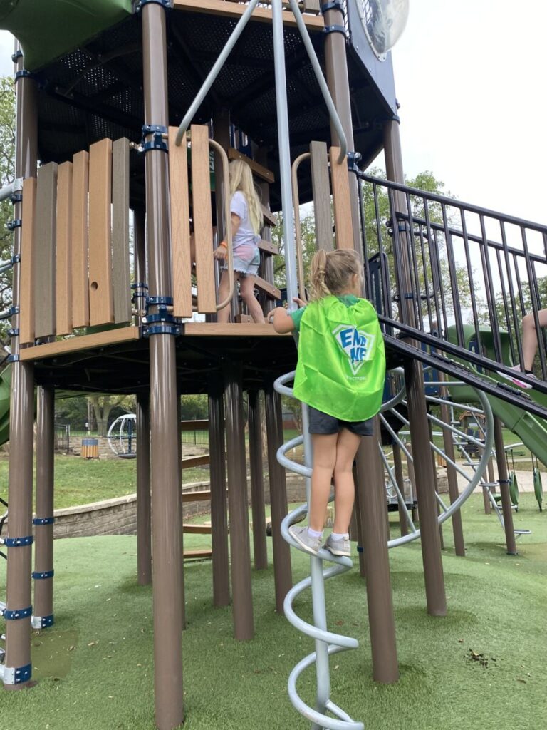 A young girl in a green cape climbs a spiral ladder to a playground structure while other children stand on the platform above.