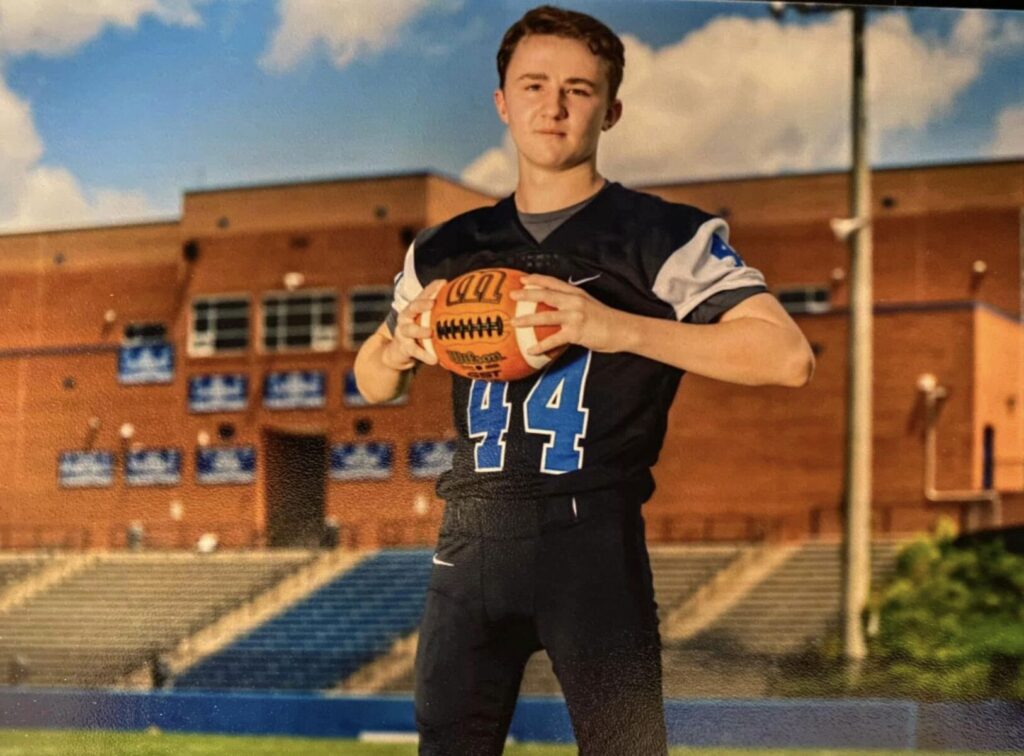 A football player in a black and blue uniform with the number 44 holds a football on a field with bleachers and a brick building in the background.