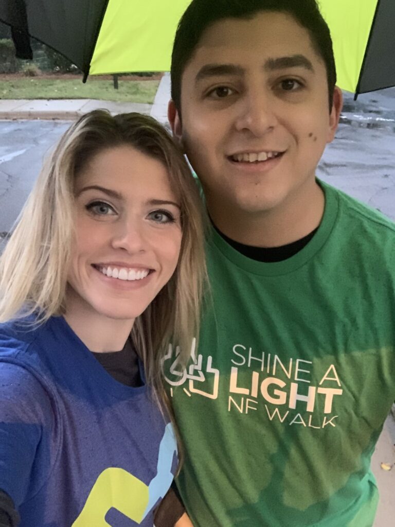 Two people stand close together under an umbrella, smiling at the camera. One wears a green "Shine a Light NF Walk" shirt; the other wears a blue shirt.