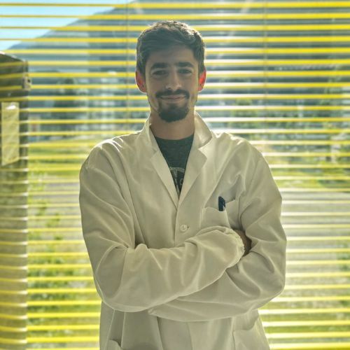 A person wearing a white lab coat stands with arms crossed in front of a window with horizontal blinds and sunlight streaming in.