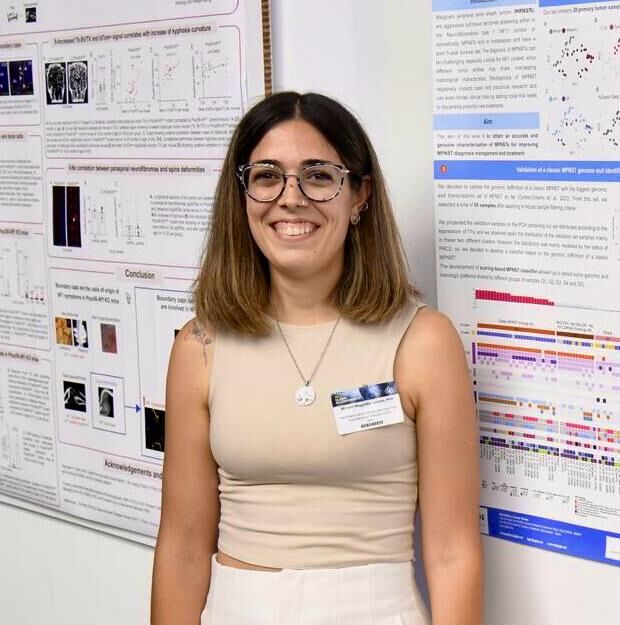 A woman wearing glasses and a sleeveless beige top stands smiling in front of scientific research posters.