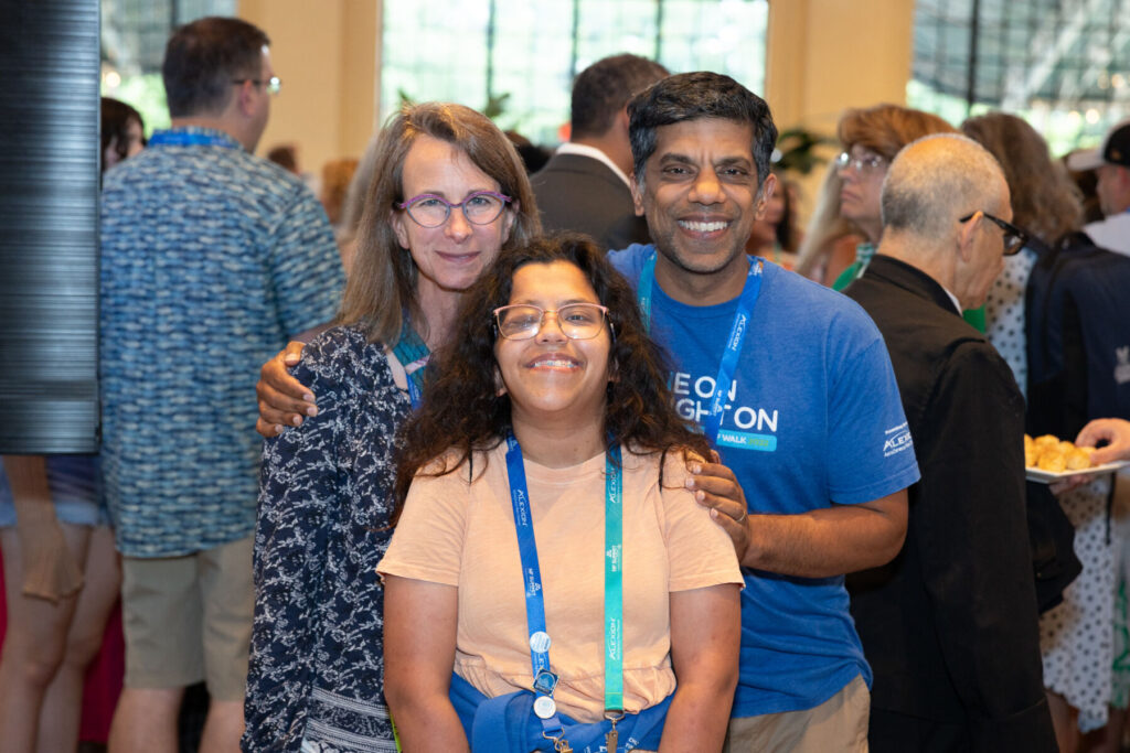 Three people are posing and smiling at an indoor event, with other attendees in the background. All are wearing event lanyards.