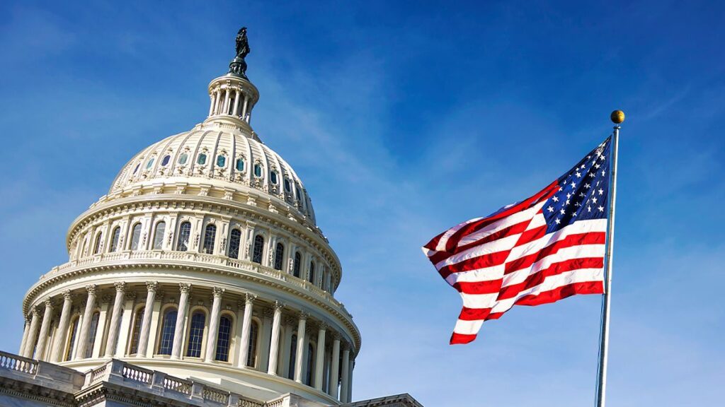 The dome of the United States Capitol building with an American flag waving on a flagpole in the foreground against a blue sky.