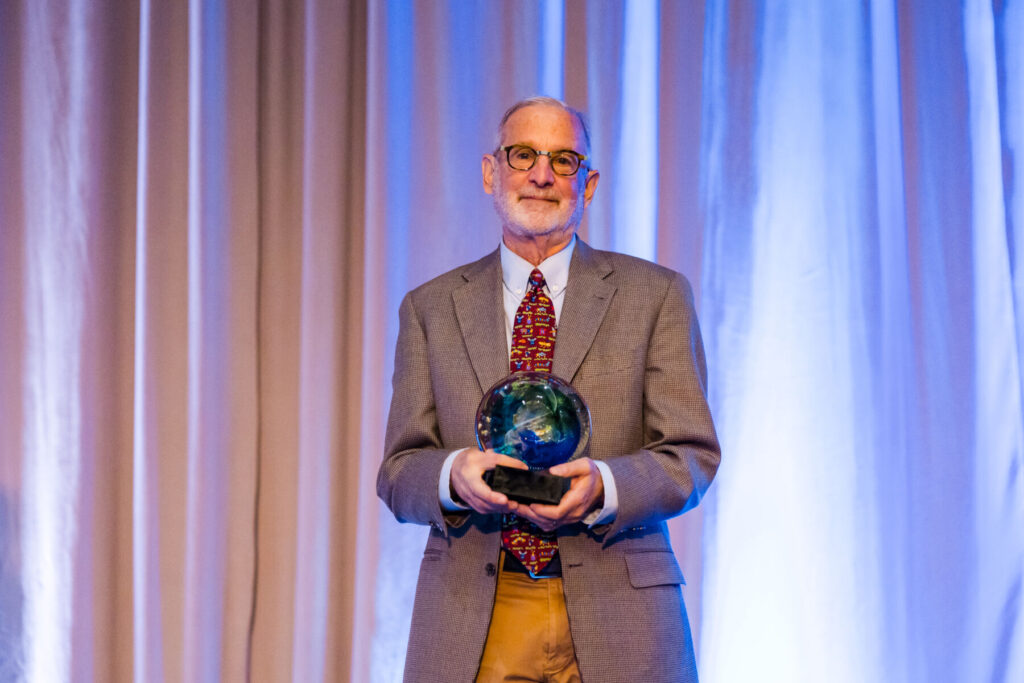 An older man in a gray suit holds a glass award while standing in front of blue and beige curtains.