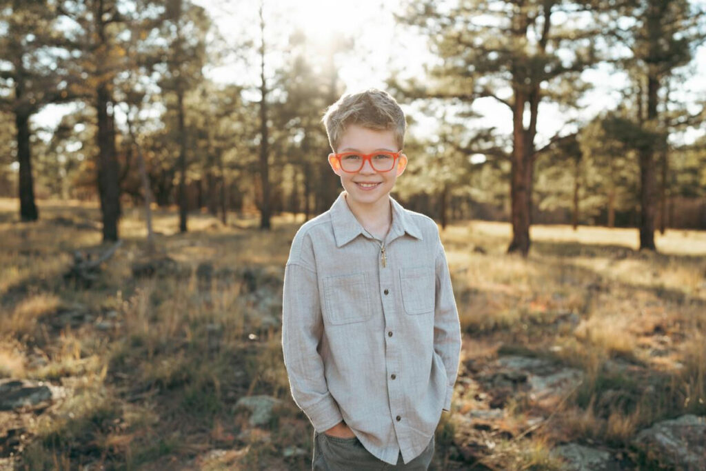 A young boy wearing red glasses and a gray shirt stands outdoors in a sunny forest clearing, smiling at the camera.